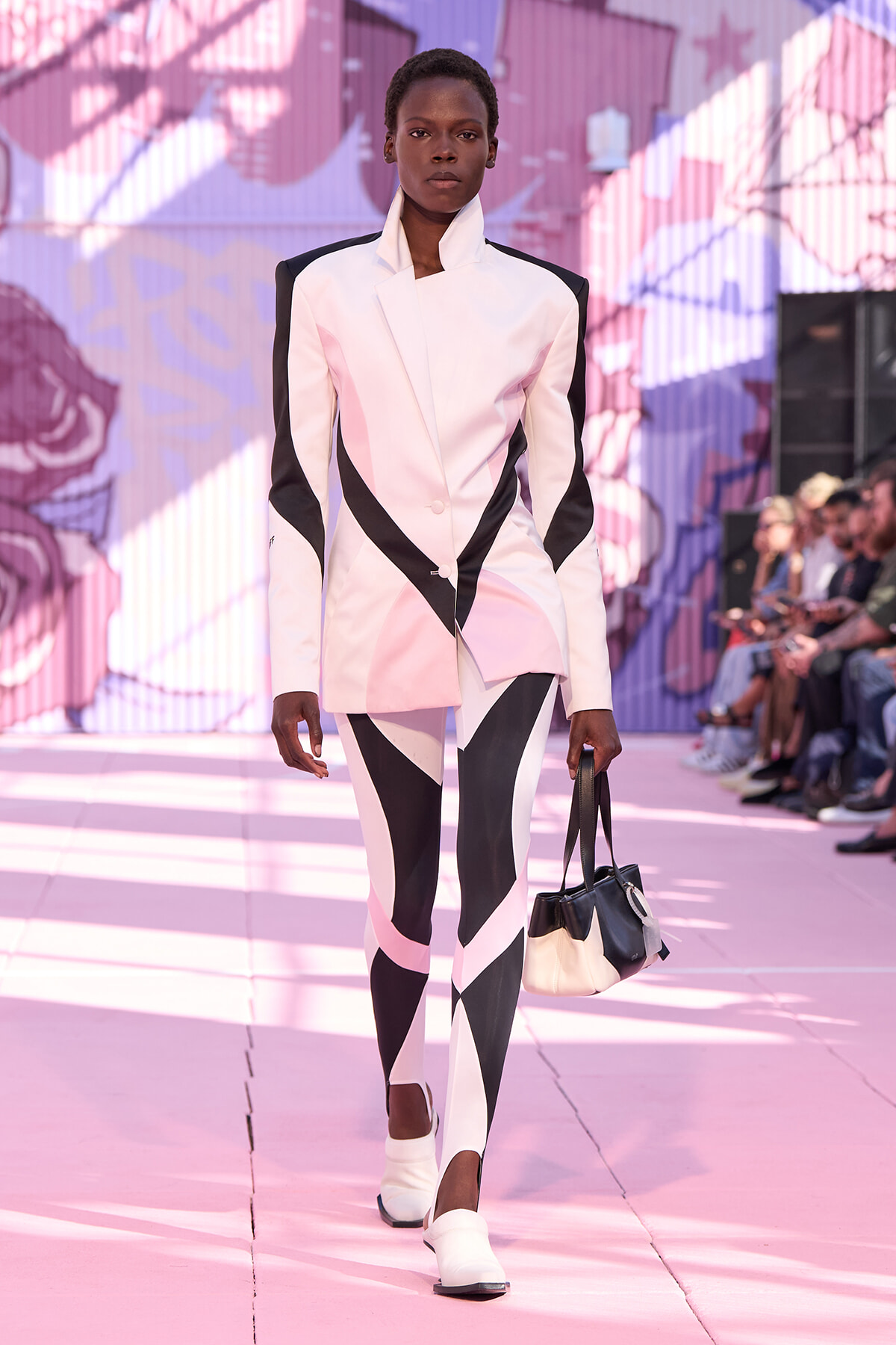 Model on a pink runway wearing a geometric pink and black blazer and leggings, carrying a black-and-cream handbag.
