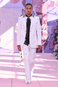 Male model walking a pink graffiti runway in a white suit with jeweled collar and black scarf/tie.