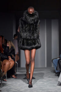 Model on a runway wearing a long, black fur coat that reaches mid-thigh, covering her neck and upper body.