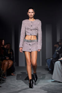Model walks the runway in a pink tweed cropped jacket and matching mini skirt, with a wide belt and black ankle boots.
