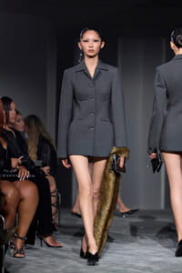 Model walks a runway in a structured gray blazer dress, holding a fur stole and a small clutch, with audience seated along the sides.