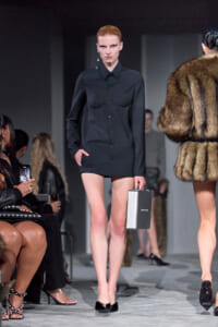 Model on a fashion runway wearing a black button-down shirt and matching shorts, holding a small silver rectangular clutch; fur-clad model nearby in the background.