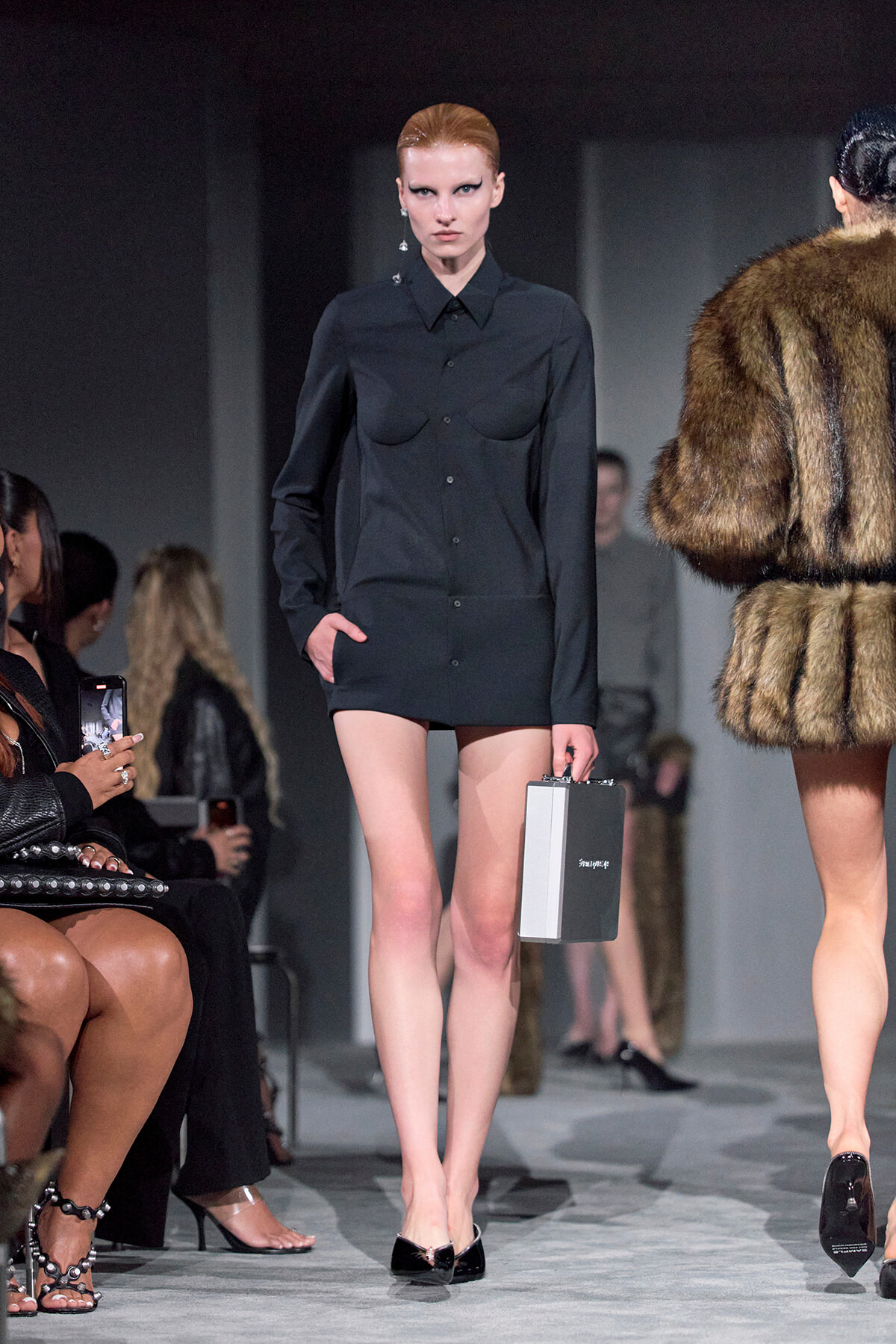 Model on a fashion runway wearing a black button-down shirt and matching shorts, holding a small silver rectangular clutch; fur-clad model nearby in the background.