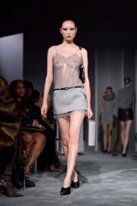Model walks a fashion runway in a sheer taupe top and light gray mini skirt, carrying a small silver box clutch and wearing black heels with a belt.