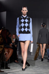Model walking a fashion show in a light blue shirt paired with a black and blue argyle sweater vest, carrying a silver rectangular clutch.