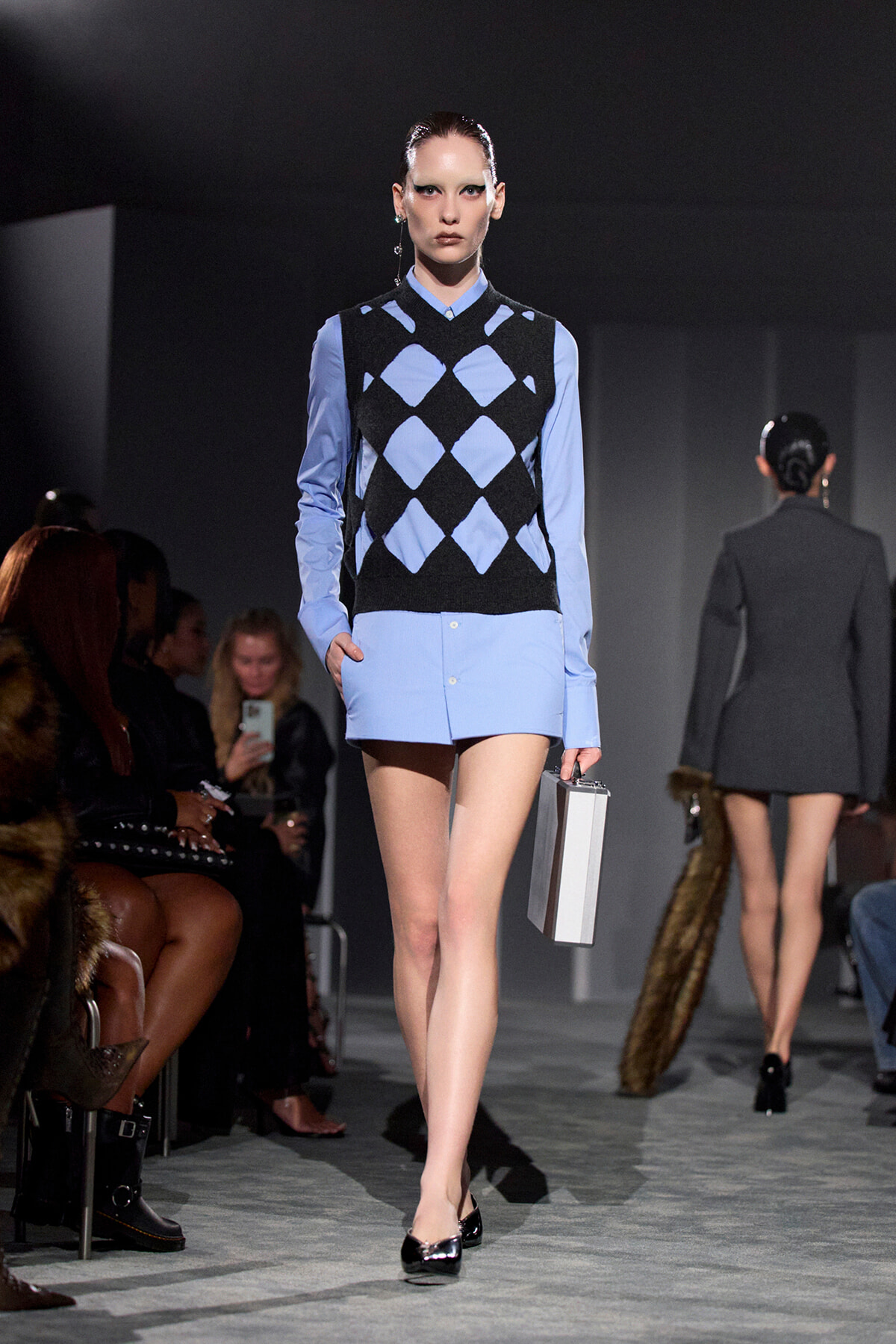 Model walking a fashion show in a light blue shirt paired with a black and blue argyle sweater vest, carrying a silver rectangular clutch.