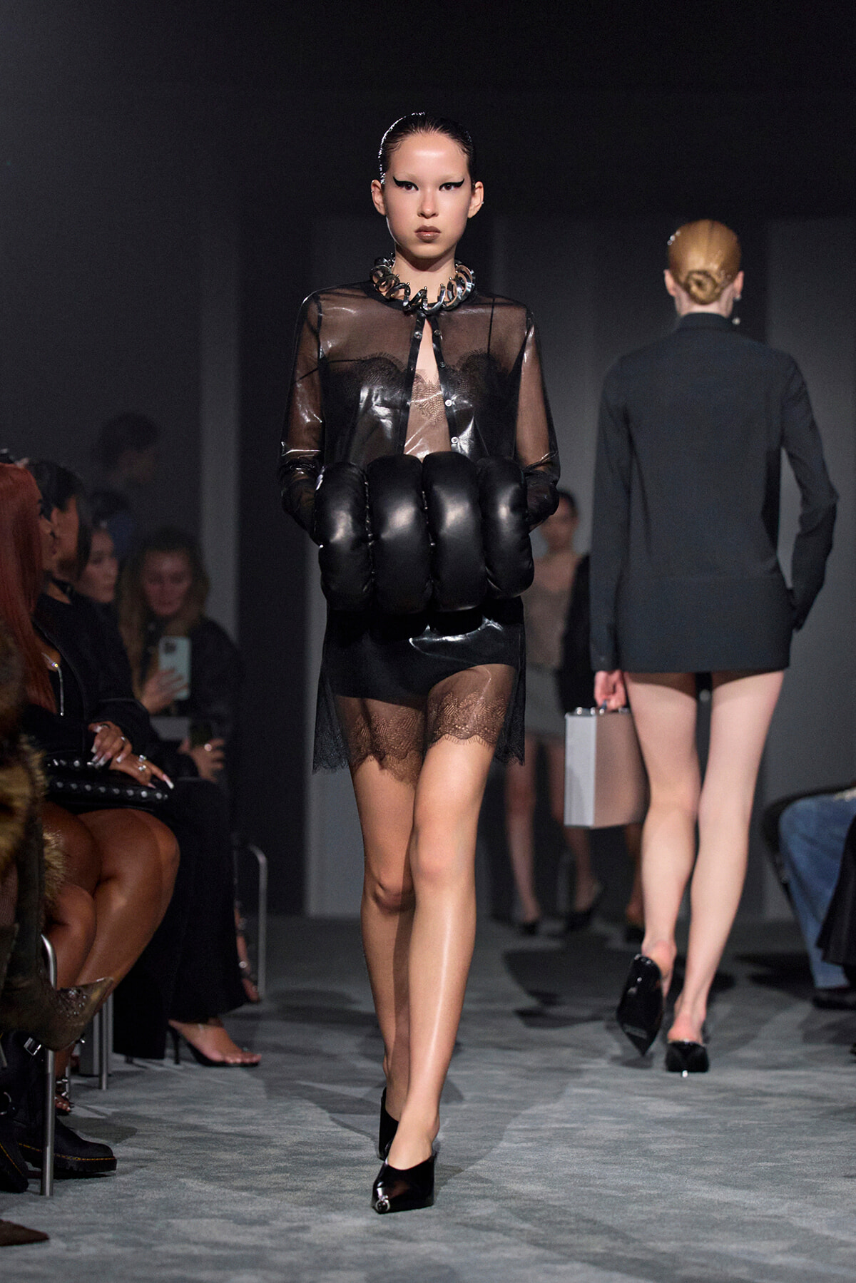Model walks the runway in a sheer black blouse, oversized black gloves, and a lace-trim mini skirt with black heels.