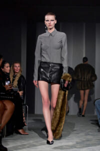 Runway model wearing a gray button-down top with a padded chest, black leather shorts, and holding a fur coat while walking the catwalk.