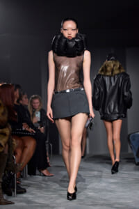 Model on runway wearing sheer top, dark mini skirt, and oversized black padded neck accessory; audience watches from sides.