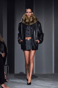 Model on runway wearing a black leather jacket with a bulky fur collar over a gray cropped suit and black heels.