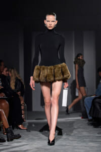 Model on a runway in a black turtleneck dress with a brown fur mini-skirt, carrying a white rectangular clutch.