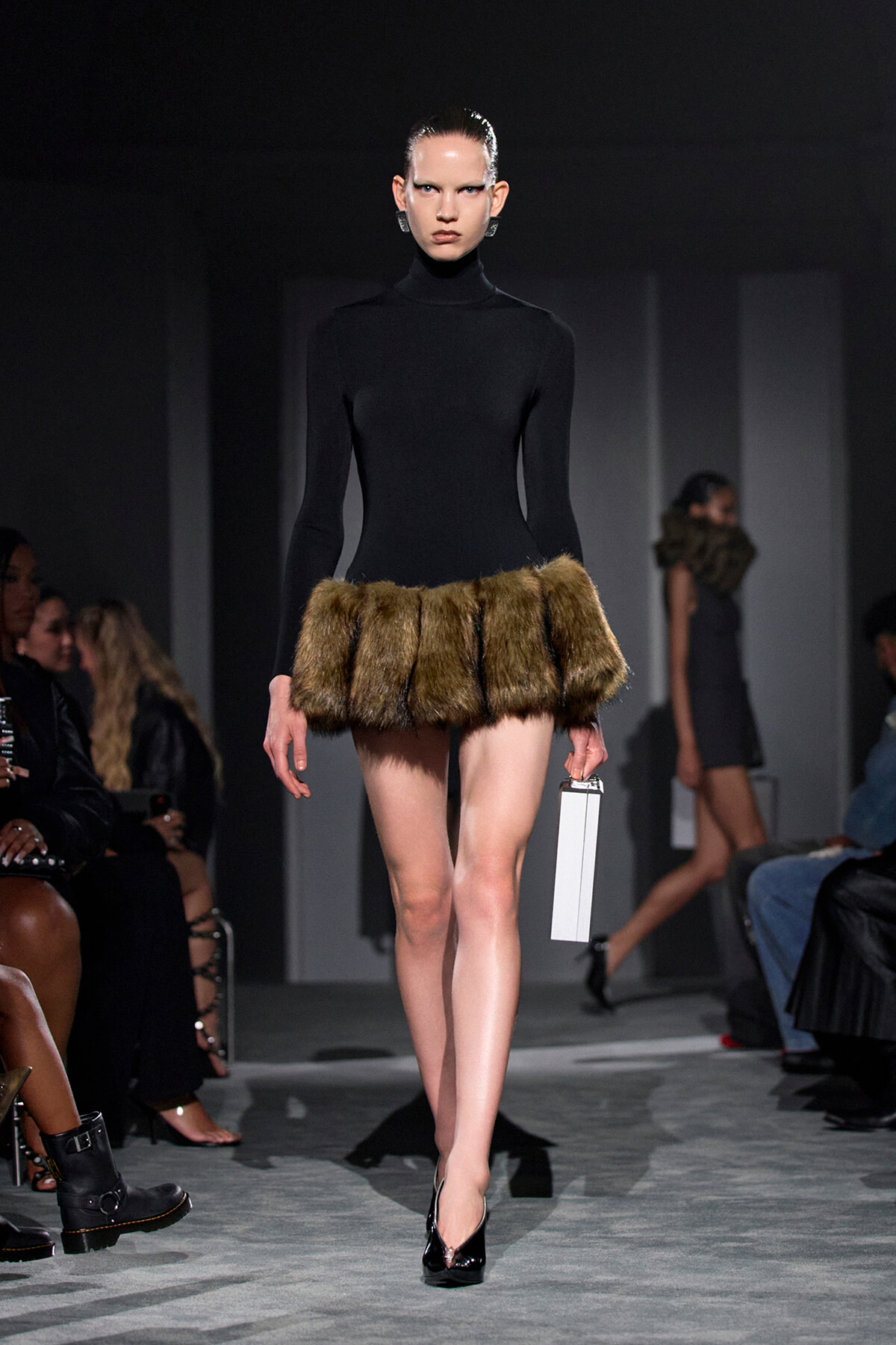Model on a runway in a black turtleneck dress with a brown fur mini-skirt, carrying a white rectangular clutch.