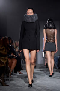 Runway model in a black tailored coat with a large beaded collar walks the catwalk; audience seated on both sides.