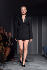Model on a runway wearing a black tailored blazer-style mini dress with satin lapels and black heels.