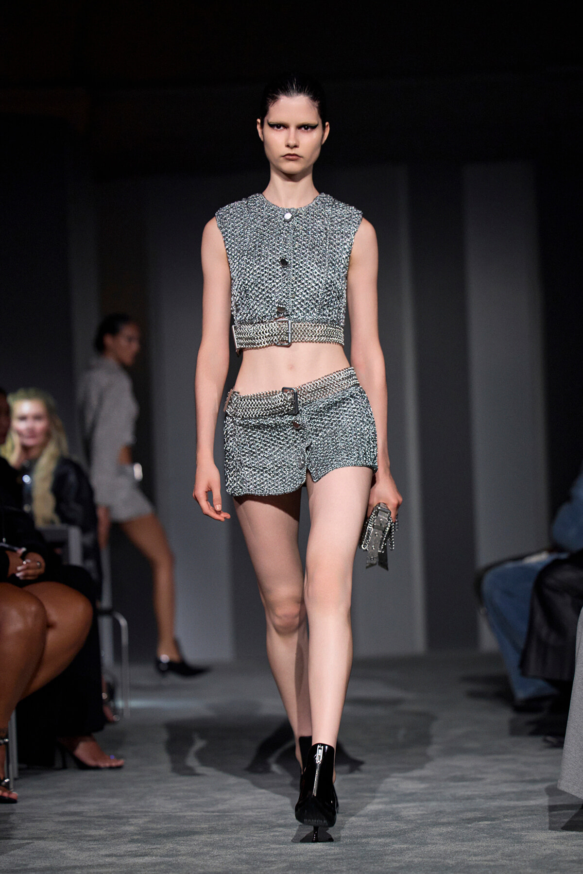 Model walks the runway in a silver textured sleeveless crop top and matching high-waisted shorts with a belt, holding a metallic clutch.