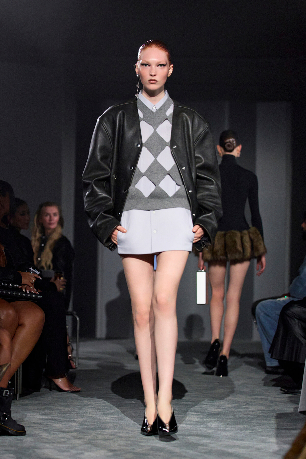 Model walking the runway in an oversized black leather jacket over a gray argyle sweater and pale mini skirt, with black heels.
