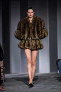 Model walks the runway in a thick brown fur coat with a voluminous silhouette and black heels, hair slicked back.