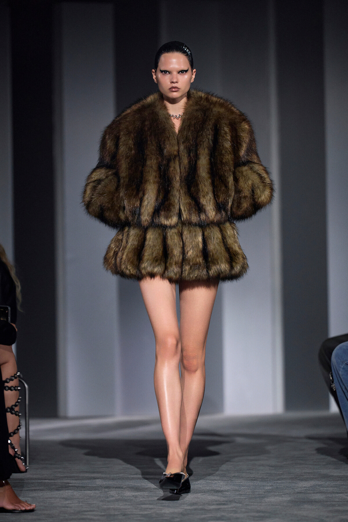 Model walks the runway in a thick brown fur coat with a voluminous silhouette and black heels, hair slicked back.