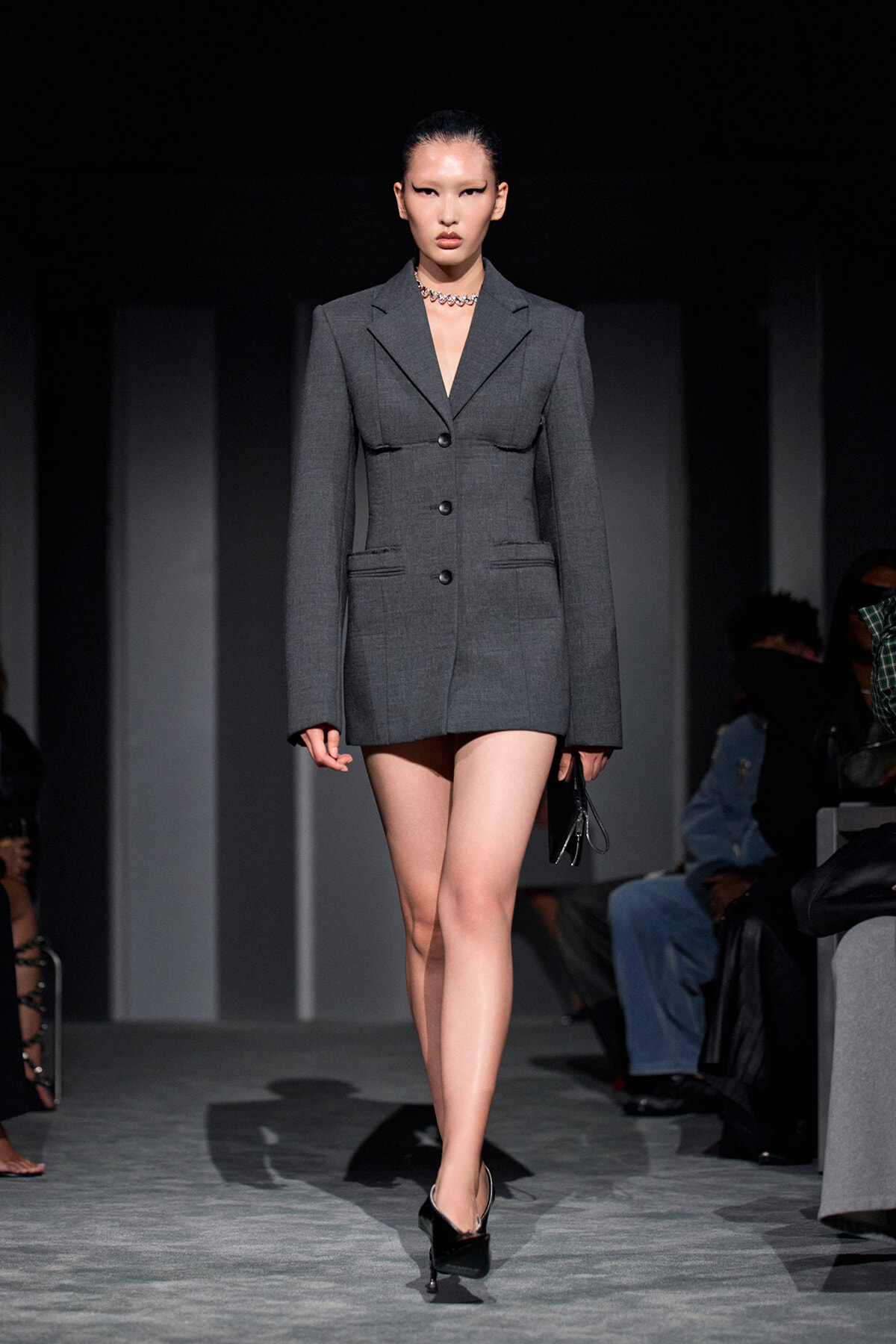 Model walking a fashion runway in a tailored gray blazer dress with black heels and a silver necklace, holding a small clutch.