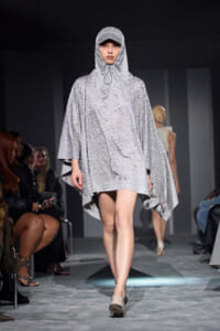 Model walking the runway in a silver, beaded poncho with hood and cap, shimmering against a dark backdrop.