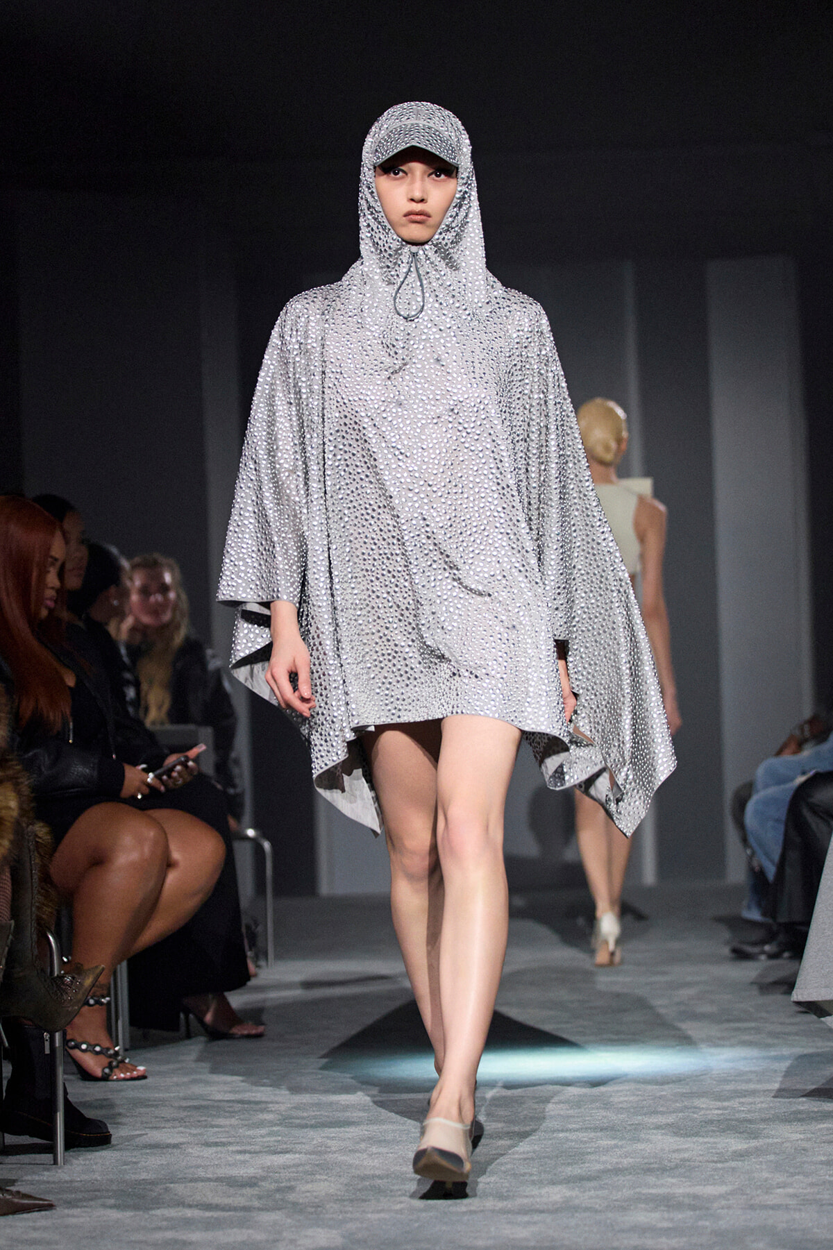 Model walking the runway in a silver, beaded poncho with hood and cap, shimmering against a dark backdrop.