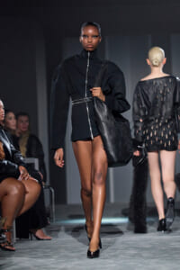 Model walks a runway in a black zip-front mini dress with an oversized jacket, carrying a large black tote.