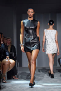 Model walks the runway in a sleeveless black leather mini dress with a front zipper and pockets.