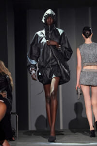 Model on a fashion-show runway wearing an oversized black hooded windbreaker with a crossbody bag; another model in a silver textured outfit walks beside her.