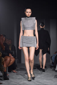 Model walks the runway in a metallic silver studded crop top and matching mini skirt; futuristic fashion with dark backdrop and audience in sides.