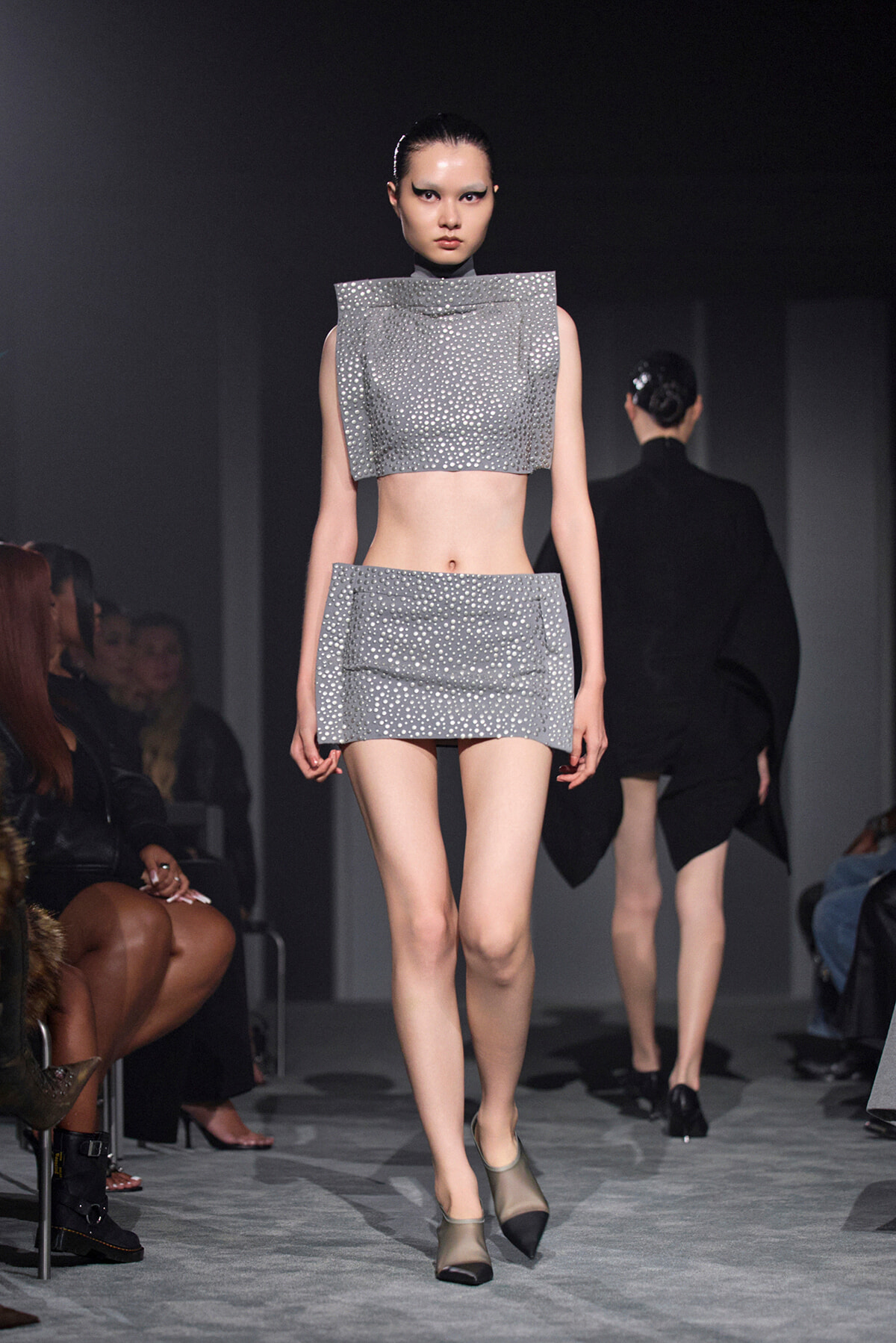 Model walks the runway in a metallic silver studded crop top and matching mini skirt; futuristic fashion with dark backdrop and audience in sides.