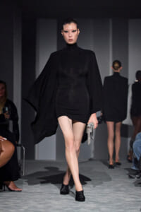Model walks the runway in a black cape-style top with a short black skirt, carrying a metallic silver clutch.
