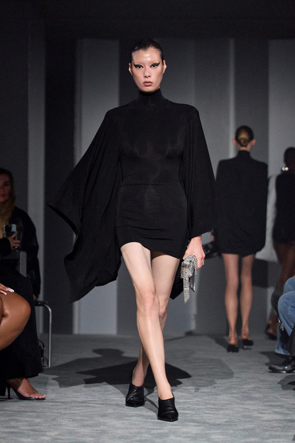 Model walks the runway in a black cape-style top with a short black skirt, carrying a metallic silver clutch.