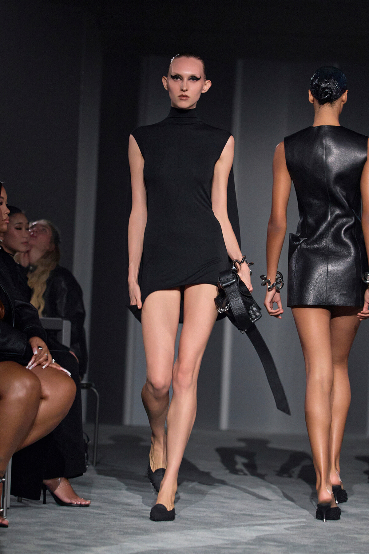 Model walks a black sleeveless mini dress with high neckline on a runway, holding a black handbag, audience on left.