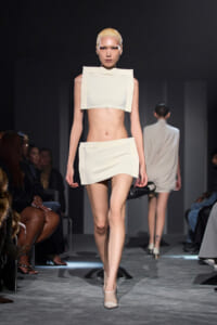 Model walks the runway in a white geometric two-piece outfit (crop top and mini skirt) with blonde slicked-back hair, audience on either side.