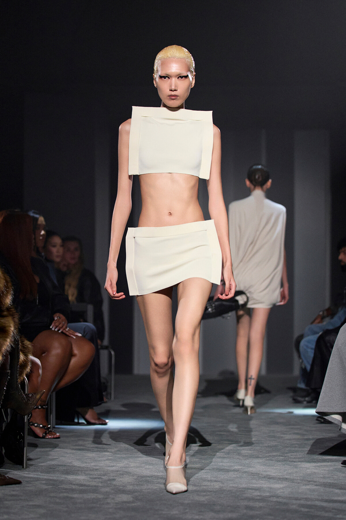 Model walks the runway in a white geometric two-piece outfit (crop top and mini skirt) with blonde slicked-back hair, audience on either side.