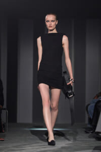Model in a sleek black sleeveless mini dress walking a fashion runway, holding a black textured clutch.