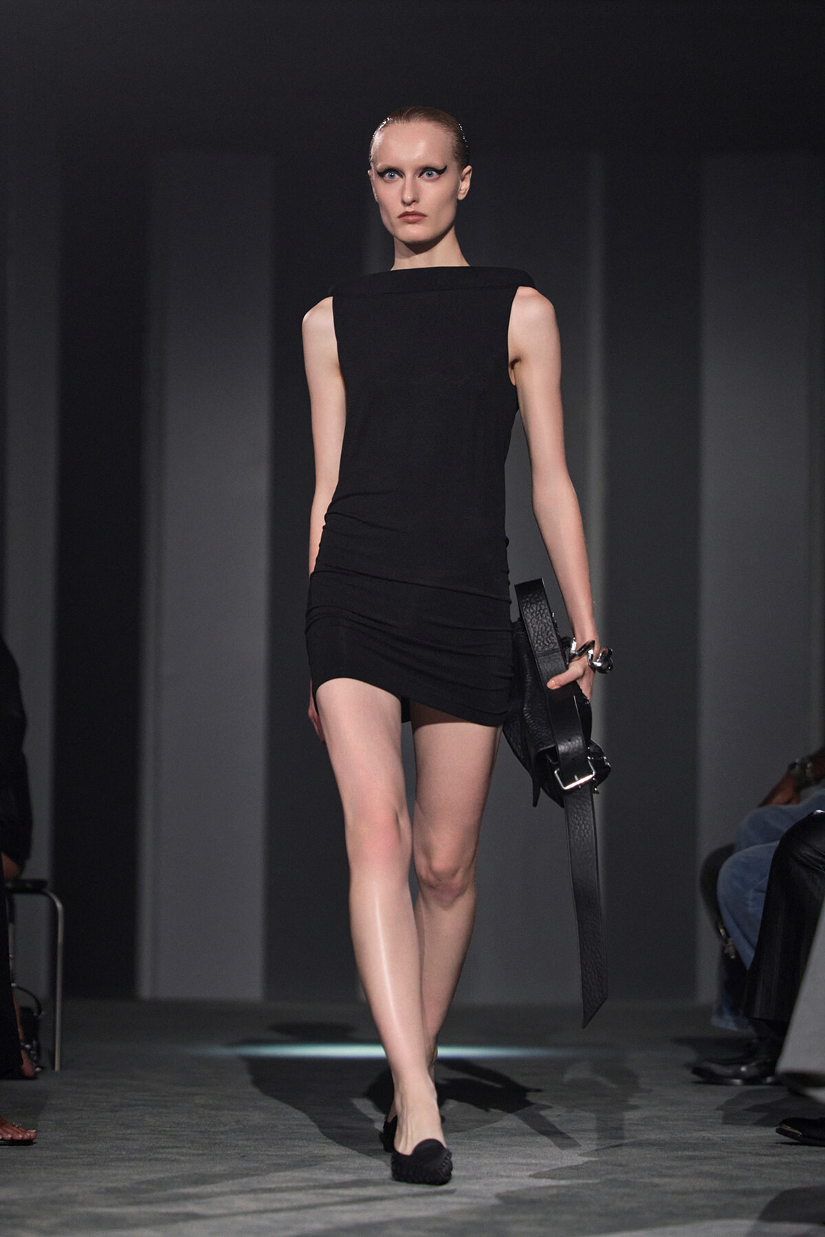 Model in a sleek black sleeveless mini dress walking a fashion runway, holding a black textured clutch.