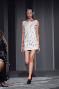 Model walks down the runway in a sleeveless white mini dress with cut-out slits and side lace trims, wearing black shoes and slicked-back hair.