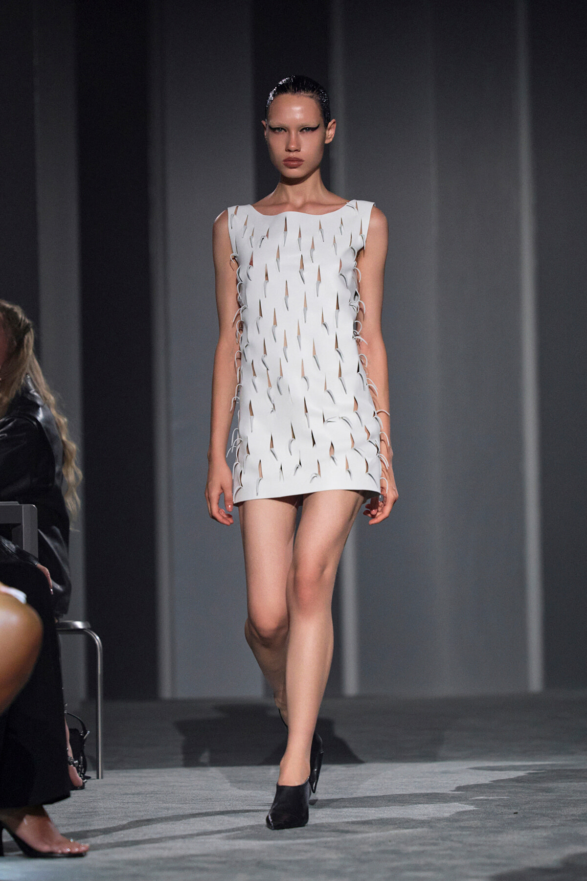 Model walks down the runway in a sleeveless white mini dress with cut-out slits and side lace trims, wearing black shoes and slicked-back hair.
