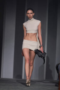 Model walks a runway in a minimalist white crop top and matching mini skirt, carrying a black embellished clutch and wearing pale heels.