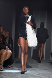 Model walks the runway in a black long-sleeve bodysuit with a large white fringed tote over her shoulder and a bold necklace, audience along the sides.