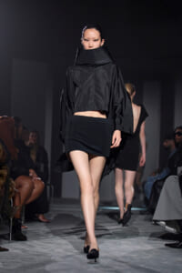 Model walks the runway in a voluminous black cape-style top with a high collar and a short black skirt, audience visible on both sides.