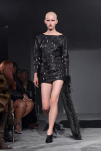 Runway model in a black mini dress with shredded cutouts and a long black fur tail accessory, walking the catwalk.