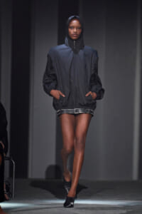 Model walking runway in a oversized black hooded windbreaker with a zip front and belt, pairing with black shorts and heeled shoes, hands in pockets.