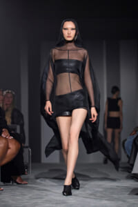 Model on a runway wearing a sheer black cape over a leather two-piece outfit with a hooded cape.