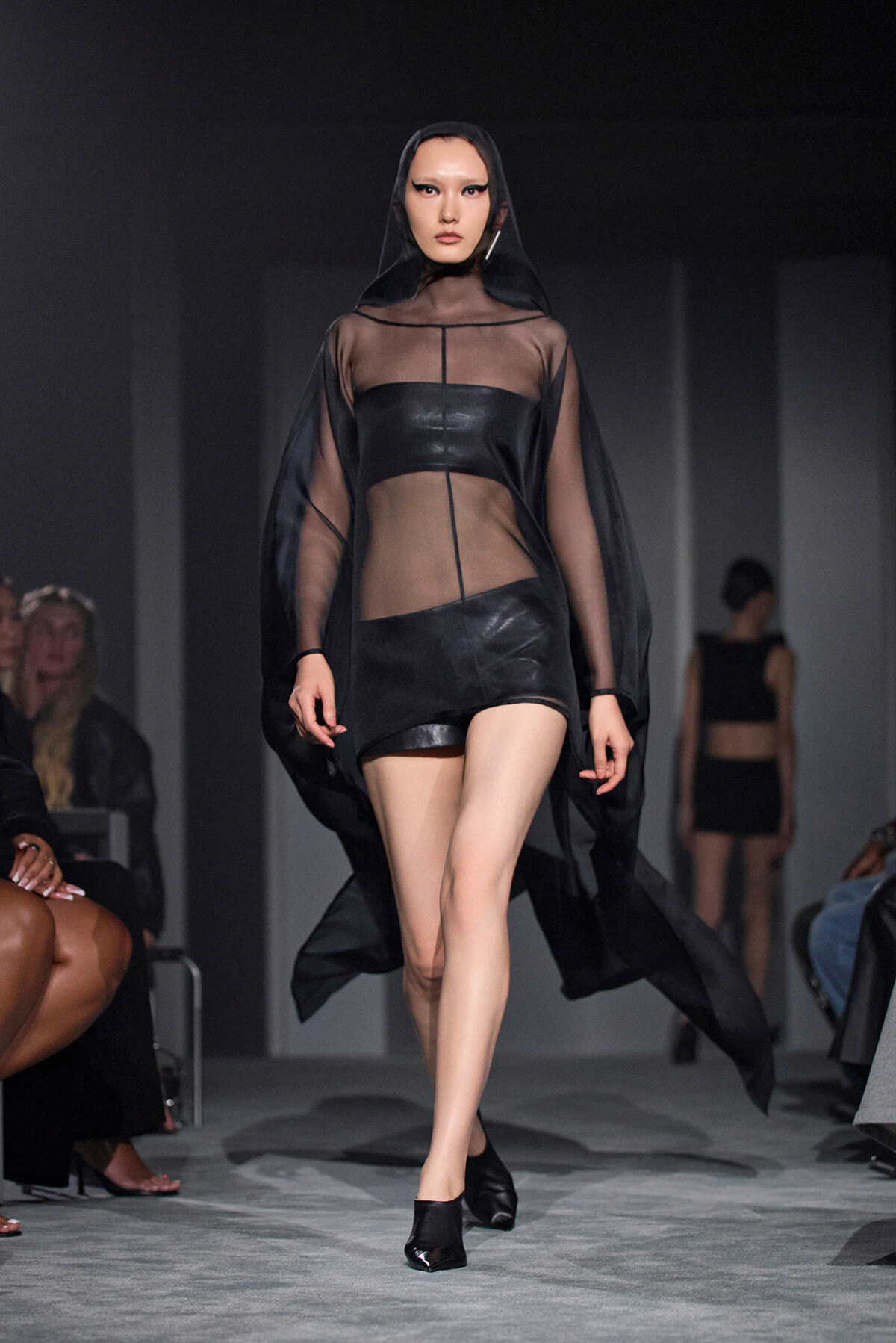 Model on a runway wearing a sheer black cape over a leather two-piece outfit with a hooded cape.
