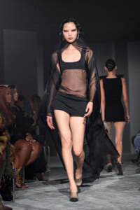 Model walking a fashion show in a sheer black cape over a black crop top and mini skirt, with seated audience along the runway.