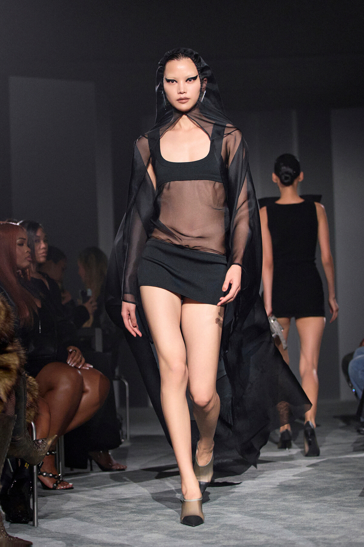 Model walking a fashion show in a sheer black cape over a black crop top and mini skirt, with seated audience along the runway.
