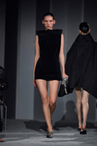Model walks the runway in a sleeveless black sculptural mini dress with a high collar, holding a silver purse.