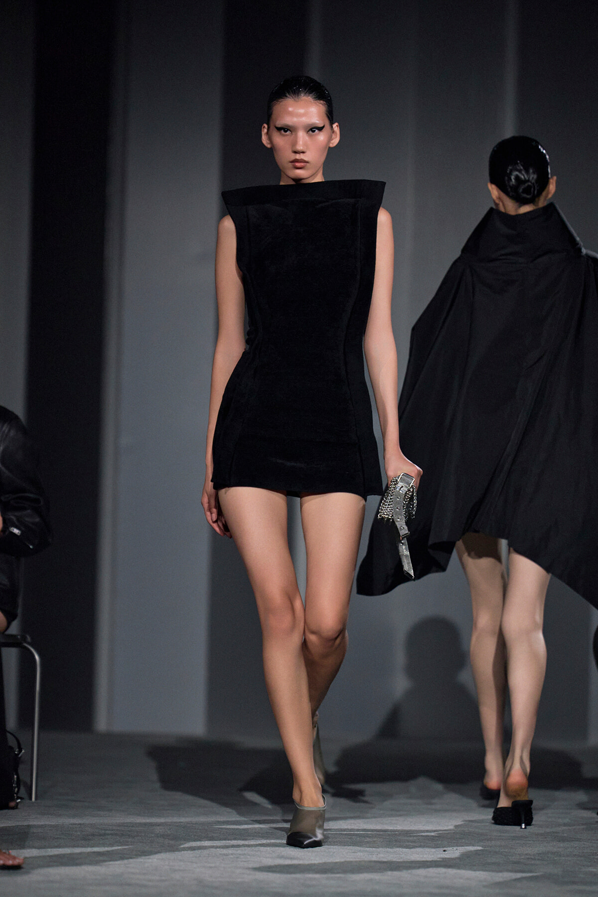 Model walks the runway in a sleeveless black sculptural mini dress with a high collar, holding a silver purse.